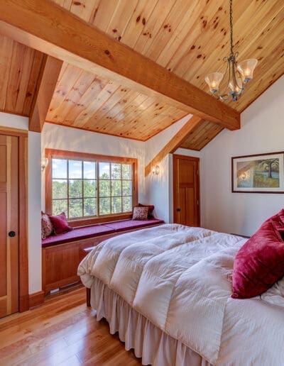 Bedroom with sloped ceiling and wood floors, featuring a window seat with red cushions.