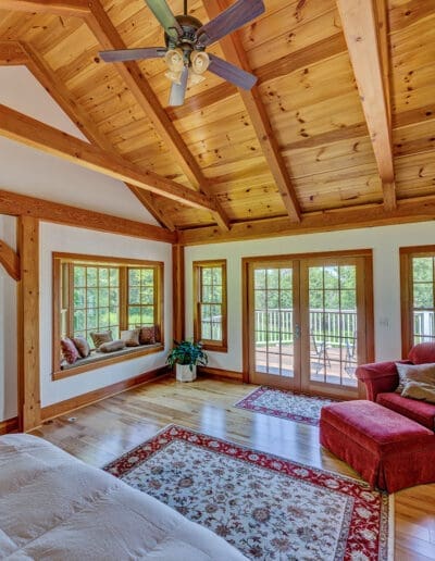 Large timber frame bedroom with tall ceilings, red stuffed arm chair, and double doors leading out to the deck.