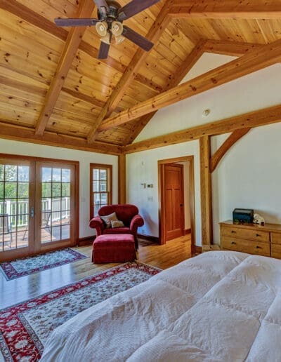 Large timber frame bedroom with tall ceilings, red stuffed arm chair, and double doors leading out to the deck.